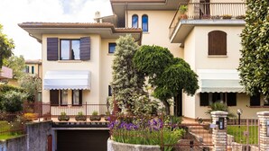 Une maison à deux étages dotée d’un porche abrité, d’une entrée fermée par un portail et d’un petit jardin agrémenté de fleurs et d’un arbre.