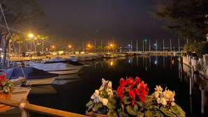 Un port de plaisance de nuit, avec des bateaux amarrés, une surface d’eau calme reflétant les lumières et une rangée de lampadaires le long du quai.