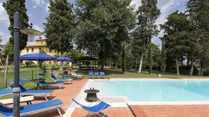 A poolside area with lounge chairs and umbrellas, surrounded by trees and a building in the background. - Belvilla by OYO Villa Santa Caterina (Cortona)