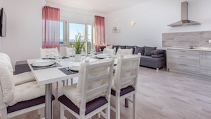 A modern dining area with a white table set for four, a grey sofa, and a kitchen area with a grey cabinet and stainless steel range hood. - Villa in Dramalj With Swimming Pool (Crikvenica)