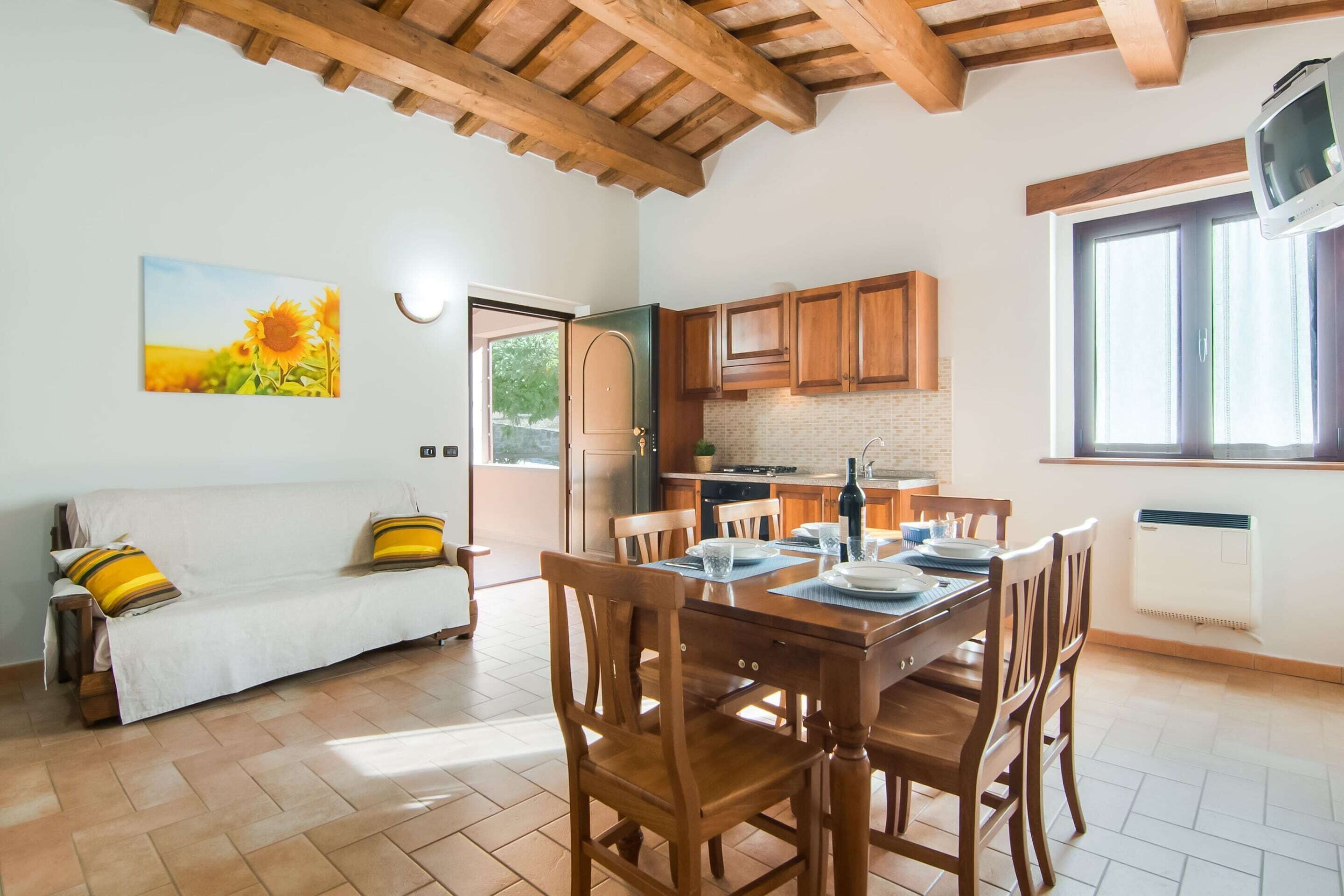 A cozy living area with a sofa, wooden dining table, and chairs, a kitchen with wooden cabinets, and a large window.