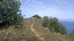 Un sentier rocailleux mène à une construction en pierre, entourée d’une clôture en bois, et bordée d’oliviers, d’où l’on peut admirer une étendue d’eau.