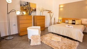 Une chambre moderne avec un grand lit, un fauteuil blanc, un tapis moelleux, une lampe sur pied et un tableau au mur.