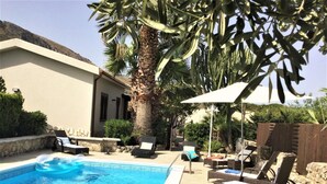Une piscine entourée de chaises longues et de parasols, bordée de palmiers et d’un mur de pierre.