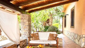 Un espace extérieur aménagé, abrité, avec un mur en pierre, un banc en bois, un canapé confortable agrémenté de coussins décoratifs, et une table sur laquelle sont disposés des fruits et des verres à vin.
