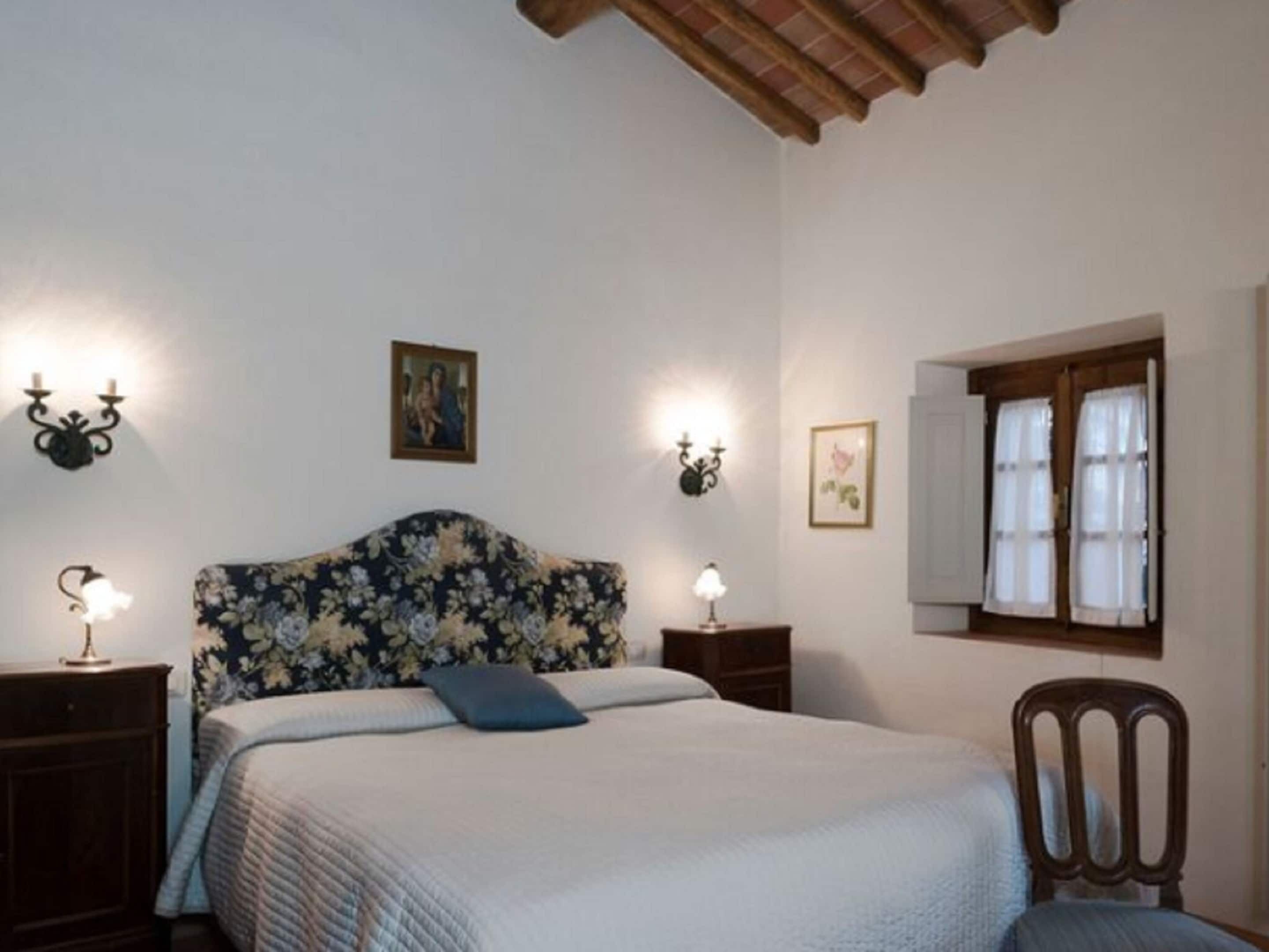 Un dormitorio con cabecera floral, una cama con almohada azul, dos mesitas de noche con lámparas, una silla de madera y un ventanal con cortinas.