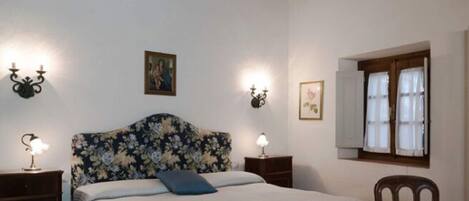 Un dormitorio con cabecera floral, una cama con almohada azul, dos mesitas de noche con lámparas, una silla de madera y un ventanal con cortinas.