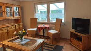 A cozy living room with a wooden cabinet, a glass-top coffee table, two wicker chairs, a small table with a red and white checkered tablecloth, and a television on a wooden stand. - Ferienhaus mit Garten in Graal-muritz (Graal Mueritz)