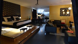 Ein modernes Hotelzimmer mit einem großen Bett, einer Sitzecke mit einer Couch und Sesseln, einem Holztisch und einem Essbereich im Hintergrund.