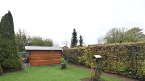 Een verzorgde tuin met een houten tuinhuisje, een vogelvoerder en een hoge naaldboom.