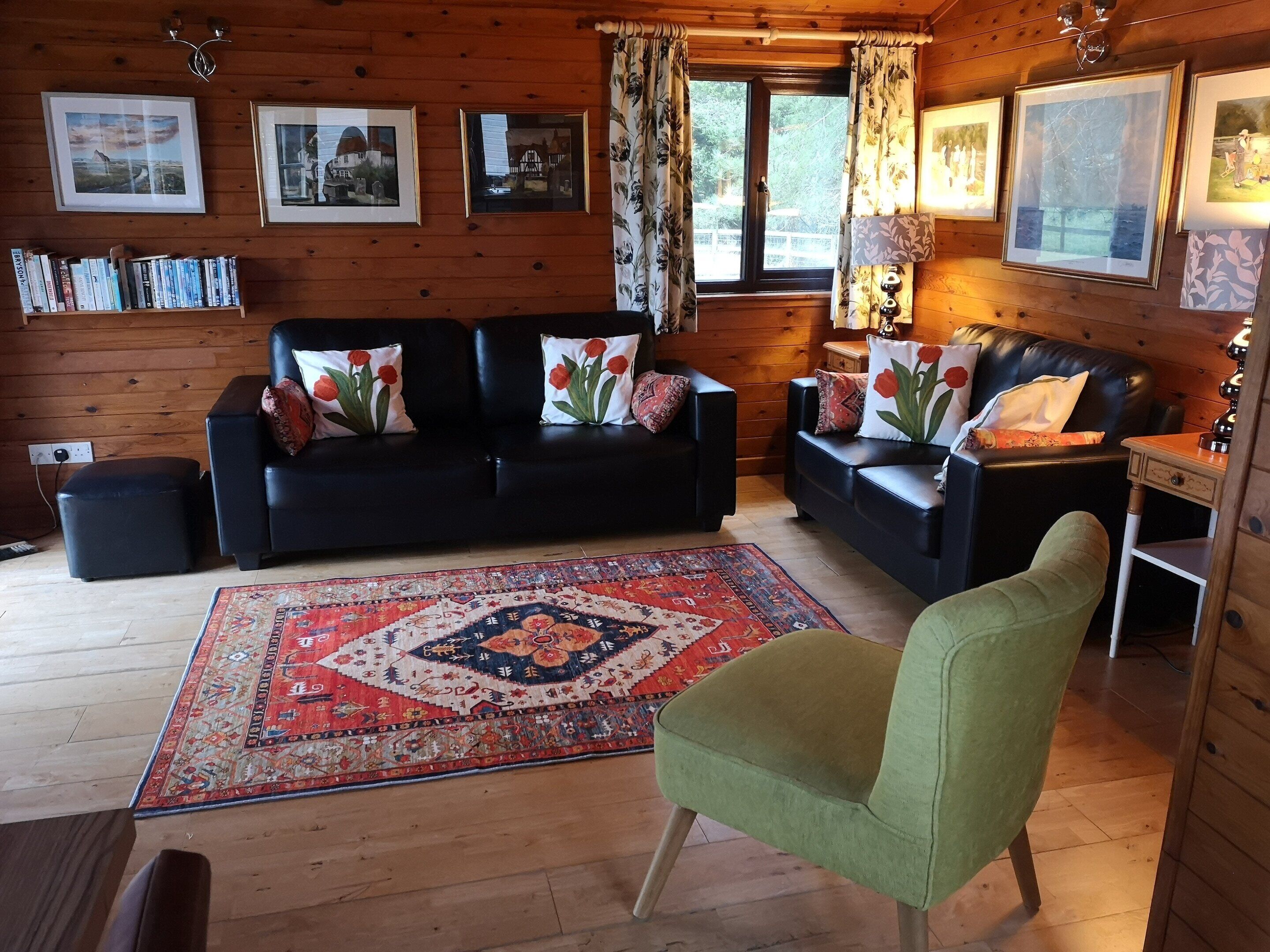 A cozy log cabin interior with two black leather sofas, a green armchair, a patterned rug, and framed pictures on the walls.