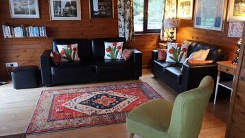 A cozy log cabin interior with two black leather sofas, a green armchair, a patterned rug, and framed pictures on the walls.