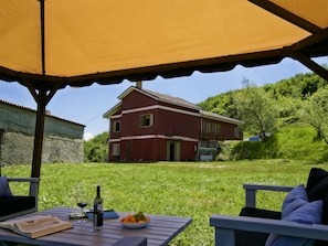 A red house with a balcony, a grassy yard, and a table with a wine bottle, glasses, and a bowl of fruit. - Spotlight Home of Corvara (Corvara)