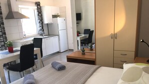 Quarto de hotel compacto com cama, escrivaninha, cadeira, mesa pequena e cozinha compacta.