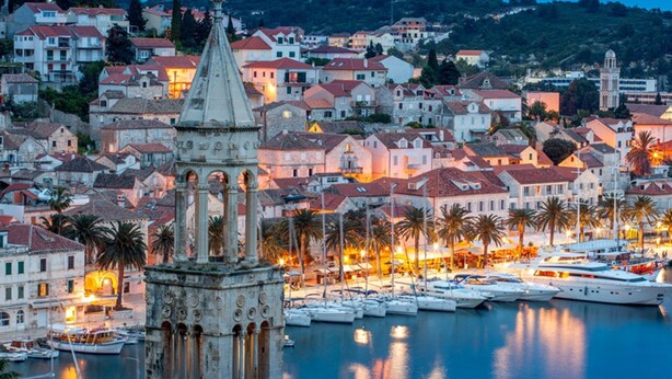 Uma vila costeira com arquitetura histórica, incluindo uma torre imponente e uma marina com barcos.