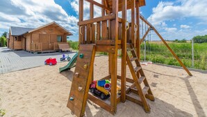 A wooden play structure with a slide and climbing wall, a small wooden building, and a sandy area with toys. - Cozy Holiday Home in Mielno near Lake (Mielno)