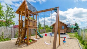 A wooden play structure with a slide and swings on a sandy area, surrounded by a wooden fence and a small building in the background. - Cozy Holiday Home in Mielno near Lake (Mielno)