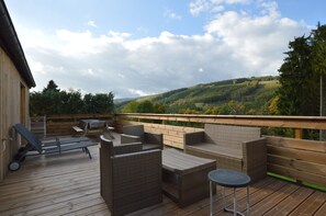 Um terraço com mobiliário de vime, uma mesa e vista para uma encosta arborizada.