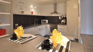 A modern kitchen with white cabinets, a black countertop, and a stainless steel range hood. There is a microwave, oven, and a small sink. A table with a striped placemat and a glass vase with yellow flowers is visible. - Countryside Holiday Home in Stoumont With Terrace, Garden (Stoumont)