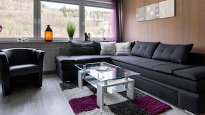 A modern living room with a black sofa, a glass coffee table, and a view of a forest through large windows. - Cozy Apartment in Willingen near Willingen Train Station (Willingen)