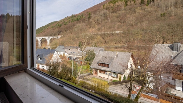 A view from a window overlooking a residential area with houses, a bridge, and a hill with trees. - Cozy Apartment in Willingen near Willingen Train Station (Willingen)