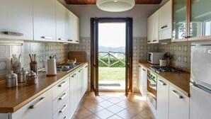 Une cuisine avec des armoires blanches, un plan de travail en bois, un luminaire suspendu et une vue sur l’extérieur par une porte ouverte.