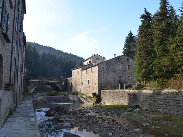 Un edificio de piedra con un puente sobre un río, rodeado de árboles y con montañas al fondo.
