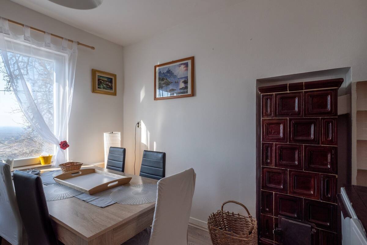 A dining area with a table, chairs, a laptop, a basket, and a window with sheer curtains.