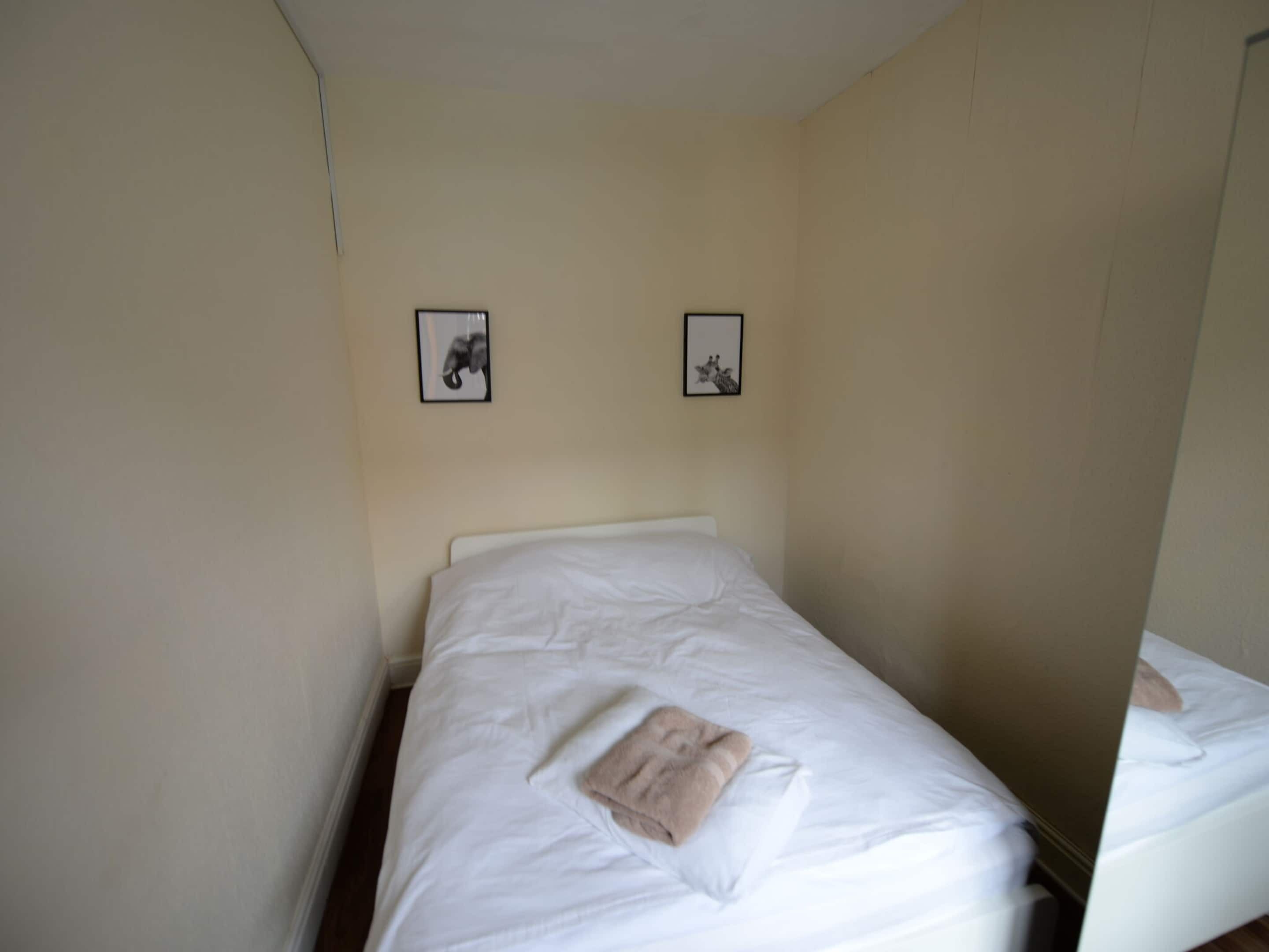 Una cama individual con ropa de cama blanca, una pared color beige y dos cuadros enmarcados.