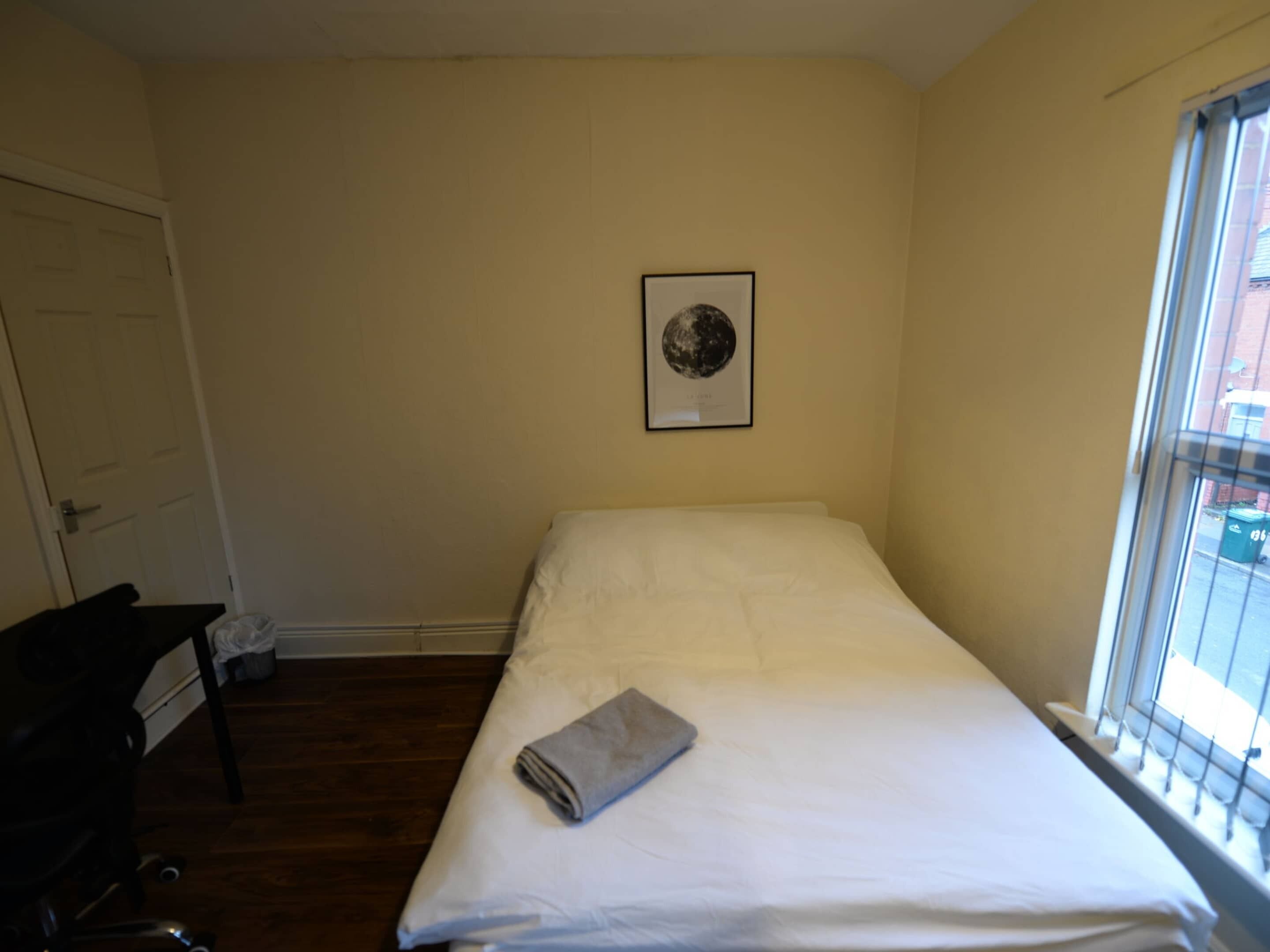 Une petite chambre avec un lit simple, un mur uni, un tableau encadré et une fenêtre.