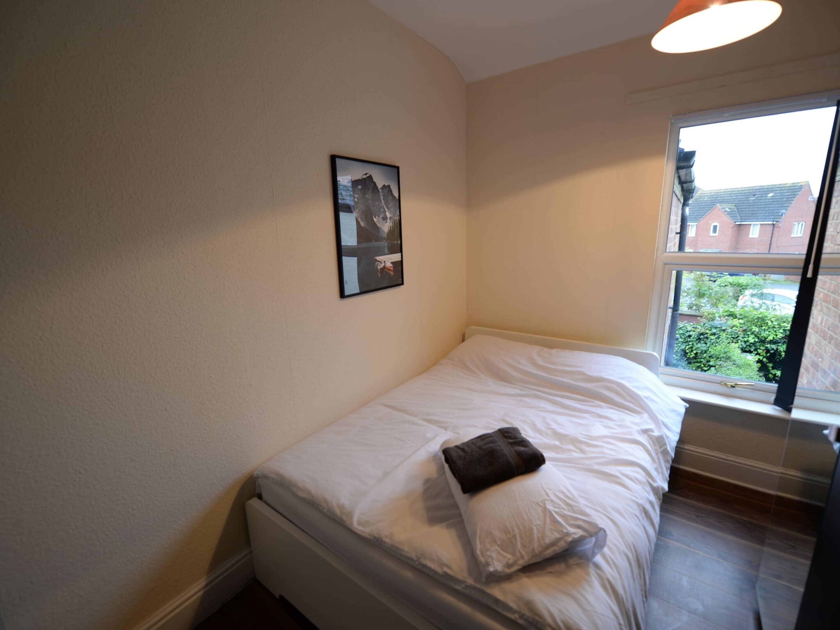 A small, neatly made bed with a white mattress and a brown pillow, a framed picture on the wall, and a window with a view of buildings and greenery outside.