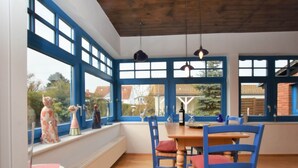 A dining area with a wooden table and blue chairs, large windows with blue frames, and decorative figurines on the windowsill. - Holiday Home With Terrace in Graal-muritz (Graal Mueritz)