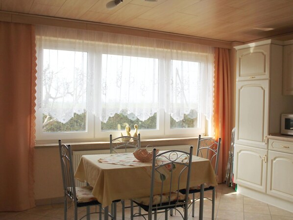 A dining area with a table set for four, a window with curtains, and a kitchen area with cabinets and a microwave. - Rustic Escape in Kropelin (Kroepelin)