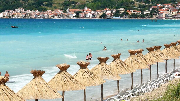 A beach with thatched umbrellas and sun loungers, a clear blue sea, and buildings in the background. - Spacious Apartment in The Croatian Islands (Malinska-Dubasnica)