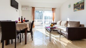A living room with a sofa, a dining table with chairs, a TV mounted on the wall, and a window with curtains. - Spacious Apartment in The Croatian Islands (Malinska-Dubasnica)
