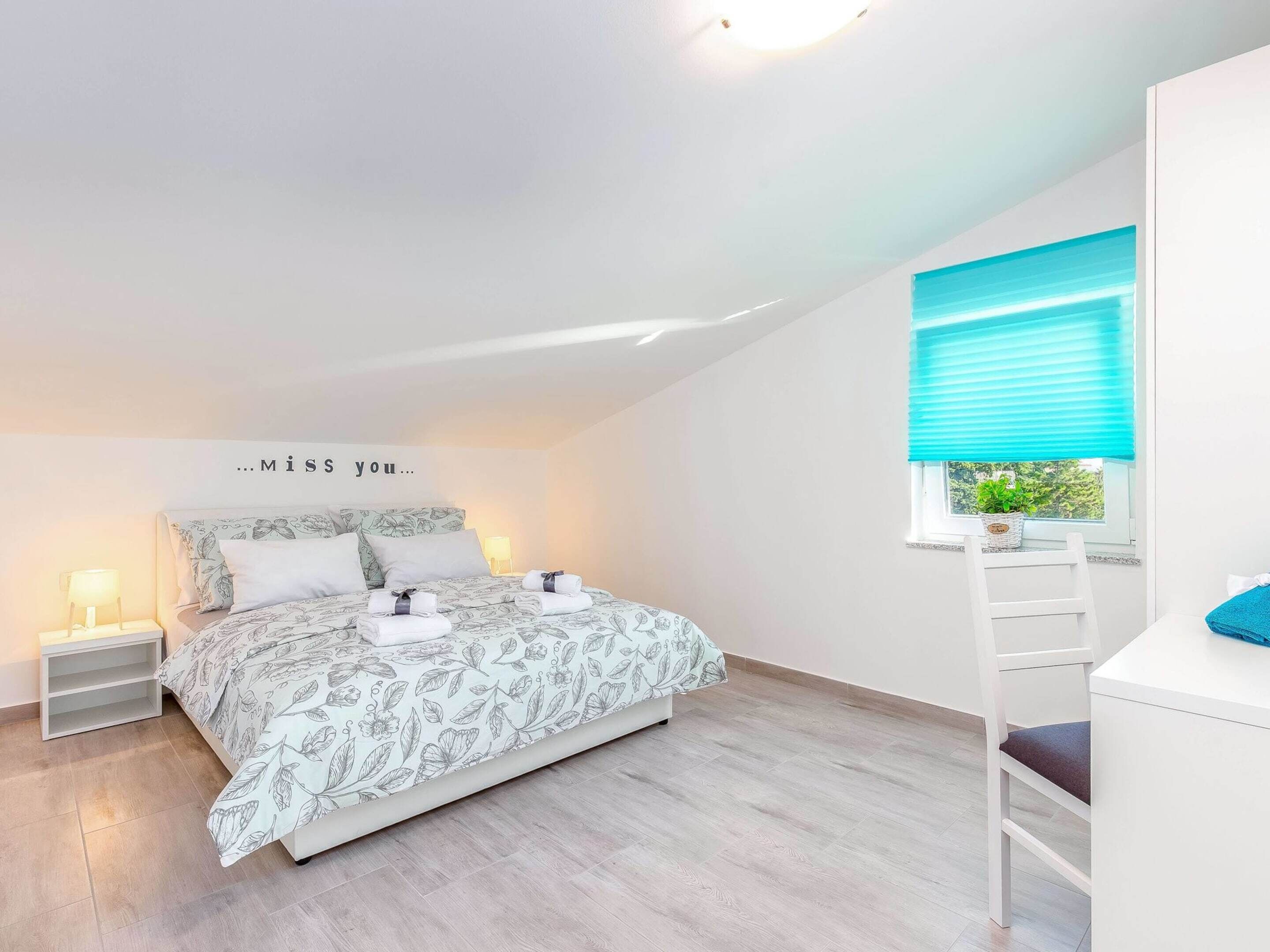 A bedroom with a bed, a nightstand, a window with blinds, and a wall-mounted shelf.