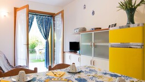 Un coin repas avec une table dressée pour quatre personnes, un meuble jaune sur lequel se trouve un téléviseur, et une vue par la fenêtre, ornée de rideaux bleus.