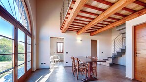 Een ruime eetruimte met een grote tafel en stoelen, houten balken in het plafond en grote ramen die zorgen voor natuurlijk licht.