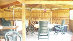 An outdoor wooden structure with a covered patio area, featuring multiple green chairs and tables, a hanging light fixture, and a stack of firewood. - Gemutliche Ferienwohnung mit Garten in Walkendorf (Walkendorf)