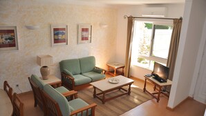 A living room with a green sofa, wooden coffee table, and a TV on a stand. There are framed pictures on the wall and a window with curtains. - Horacio Villa is Located near Center of Vilamoura (Loulé)