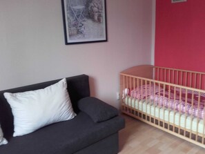 A room with a dark grey sofa, a white pillow, a crib with a checkered blanket, and a framed picture on the wall. - Eine Gemutliche Ferienwohnung in Wismar (Wismar)