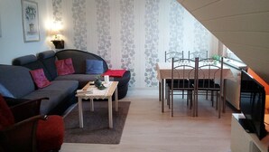Olohuone, jossa on sohva, pieni pöytä, ruokapöytä tuoleineen, televisio ja kuviollinen tapetti.