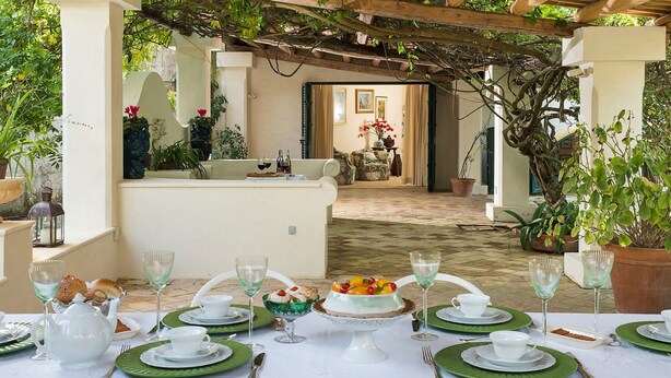 Un área de comedor al aire libre con una pérgola, muebles blancos y plantas en macetas.