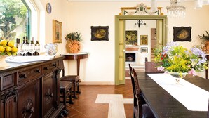 Un comedor con una mesa larga, sillas y una araña. Se observa una ventana con vista a árboles y una puerta que da a otra habitación.
