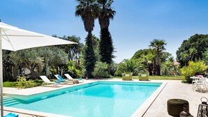 Una piscina con sillas de descanso y sombrillas, rodeada de follaje y árboles altos.