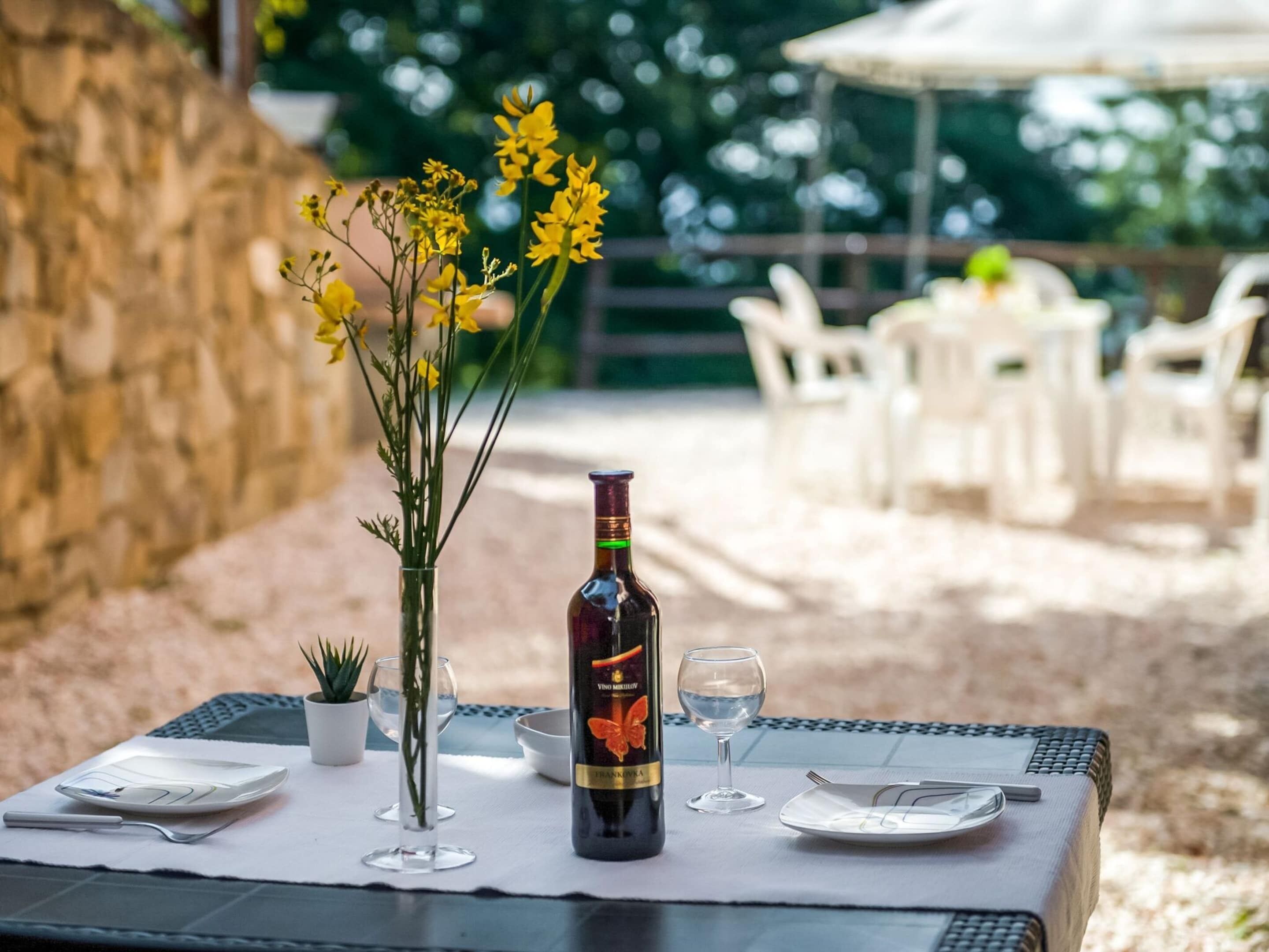 A table set for dining outdoors with a bottle of wine, glasses, and a vase of flowers.