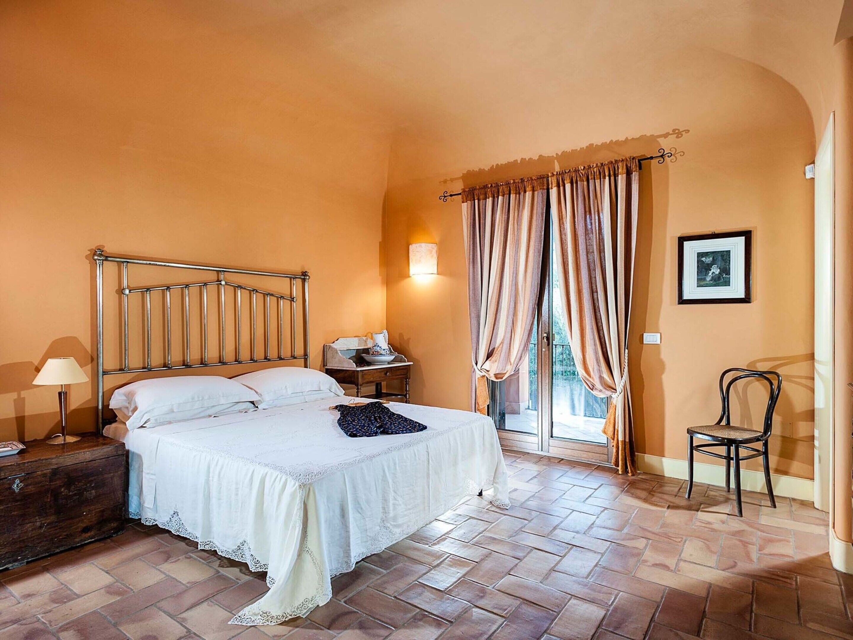 A bedroom with a metal bed frame, a bedside table with a lamp, a chair, and a view through a window with curtains.