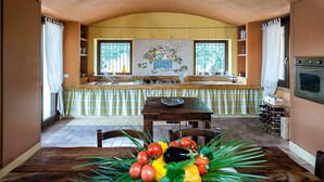 A dining area with a wooden table, chairs, and a floral centerpiece. There are two pendant lights above the table, and a kitchen area with an oven and wine rack is visible in the background. - Belvilla by OYO Villa Arangea Dieci (Marsala)