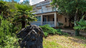 A two-story building with a balcony, surrounded by lush greenery and a large rock in the foreground. - Nice Apartment With Terrace Near the Beach (Malinska-Dubasnica)