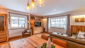 A cozy living room with a wooden dining table, a sofa, and a TV. There are windows with curtains, a fireplace, and a decorative tree on the table. - Charming House With Sauna, Jacuzzi in Bullange (Buellingen)